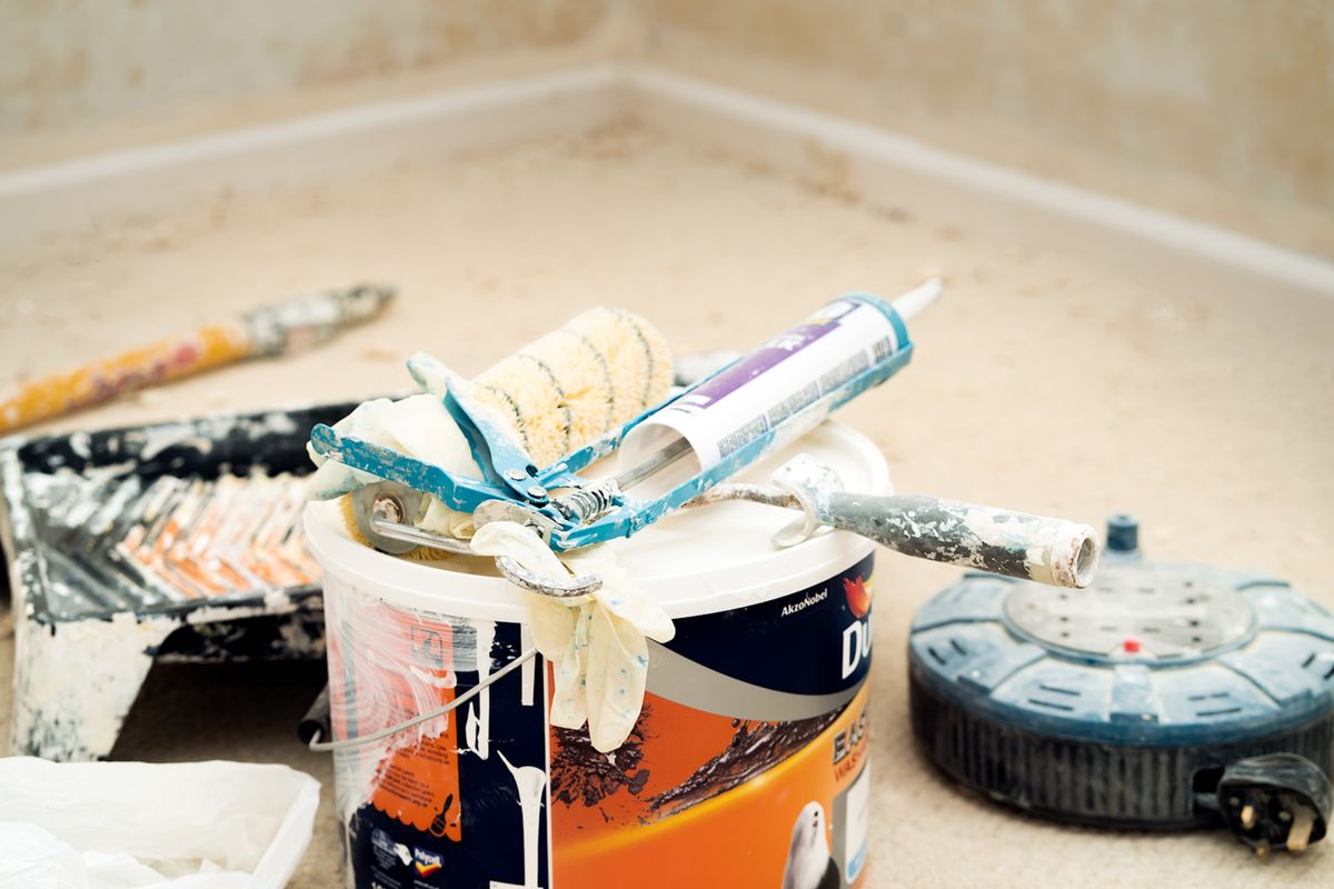 Paint bucket, roller, caulk gun, and finishing tools staged on a protected floor during ceiling work